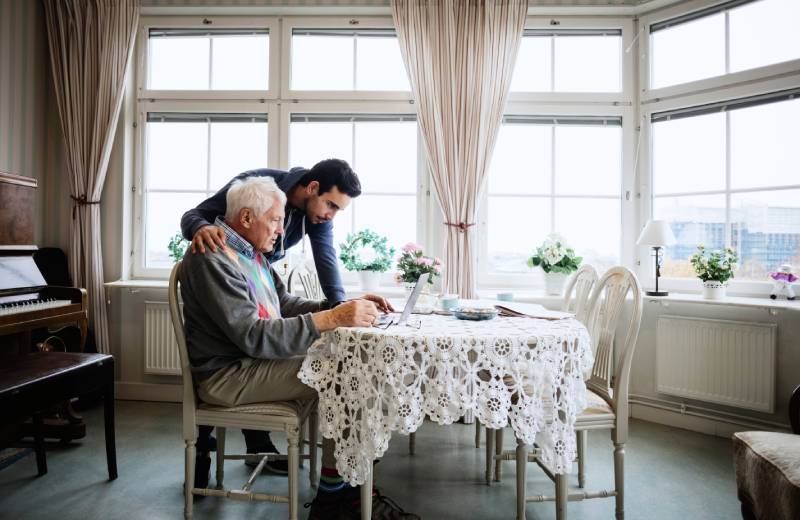 A younger man standing next to a seated older man, as they both look at a laptop